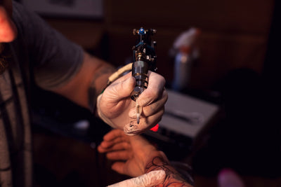 Tatuador profissional de luvas tatuando o braço de uma pessoa após explicar os cuidados com a tatuagem que o cliente deve ter após o procedimento.