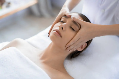 Mulher deitada em uma maca recebendo massagem facial de uma profissional.